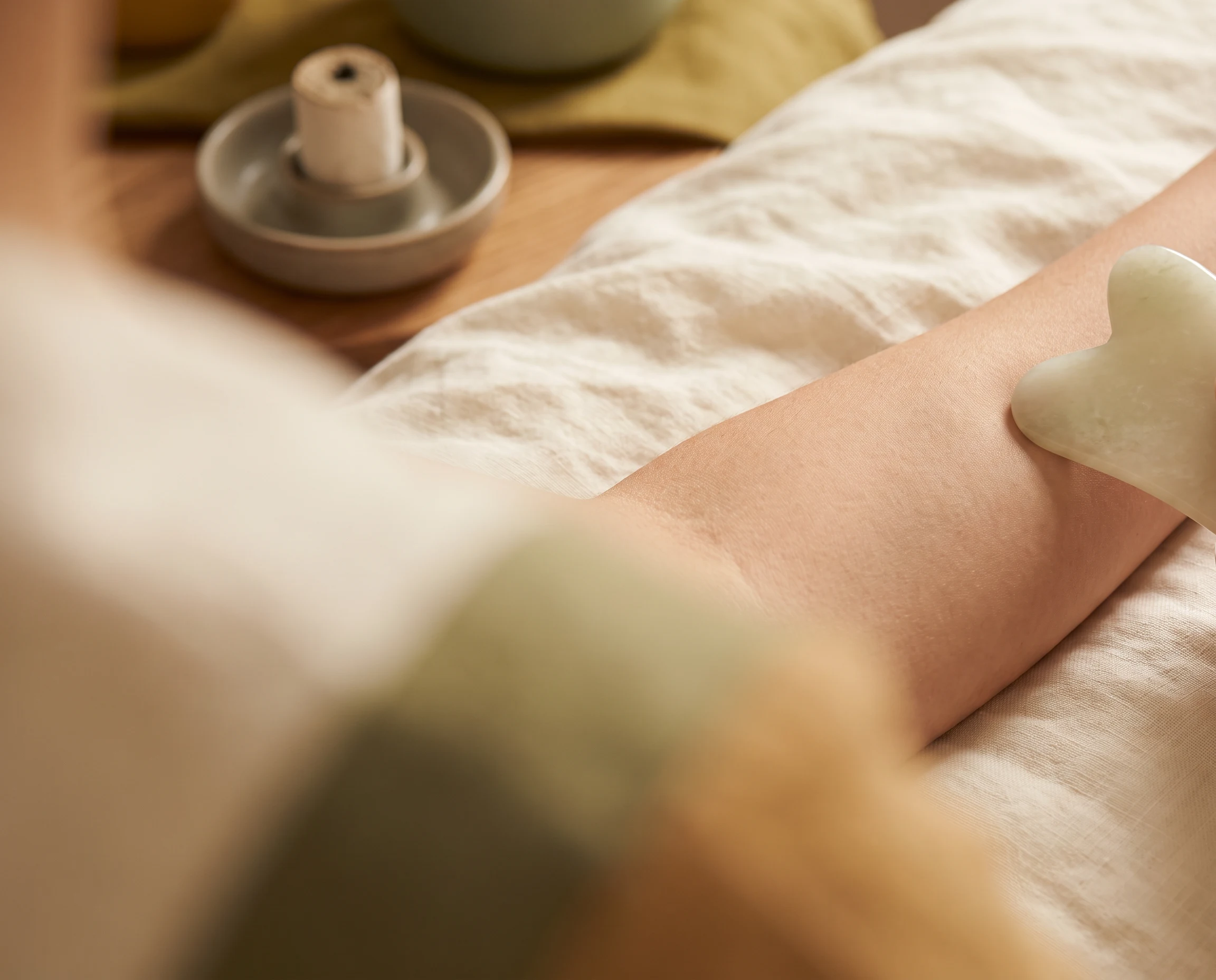 A pale jade gua sha tool resting on an anonymous forearm with moxibustion elements in the background.